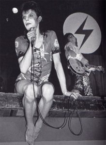 [image: Ziggy Stardust perches with mic on edge of stage, wearing a playsuit - black and white image]